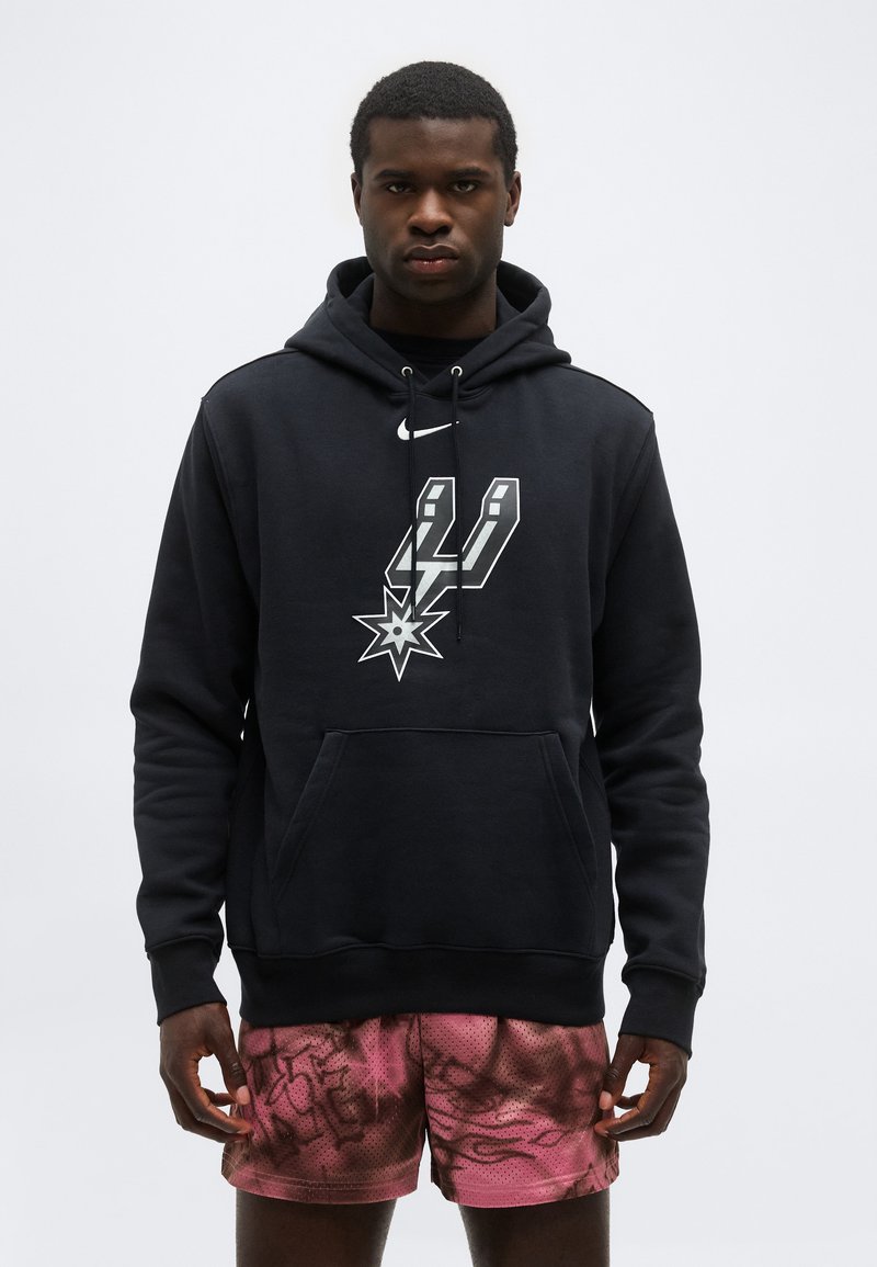 Άνδρας που φοράει μαύρο φούτερ με κουκούλα με το λογότυπο των San Antonio Spurs και ροζ σορτς με σχέδια, στέκεται μπροστά σε απλό ανοιχτό φόντο.