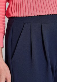 Gros plan d'une personne portant un pantalon plissé bleu marine et un pull rose côtelé, avec sa main gauche visible sur le côté.