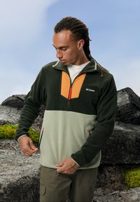 Tamnozeleni flis pulover s narančastim prednjim panelom, polu-zip zatvaračem, dva bočna džepa i crvenim akcentima. Ima teksturirani materijal i opušteni kroj.