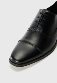 Chaussure oxford en cuir noir avec un bout pointu, une texture lisse, des coutures ton sur ton et des lacets étroits. La semelle a un léger bord pour une meilleure adhérence.