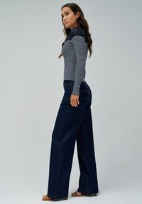 Pantalons en denim navy à jambes larges avec des coutures orange contrastantes, associés à un haut fitted à rayures noires et blanches à manches longues. Texture douce.