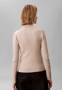 Suéter de cuello alto beige con canalé y mangas largas, con una pequeña etiqueta de la marca en la parte trasera, combinado con vaqueros de mezclilla oscuros.