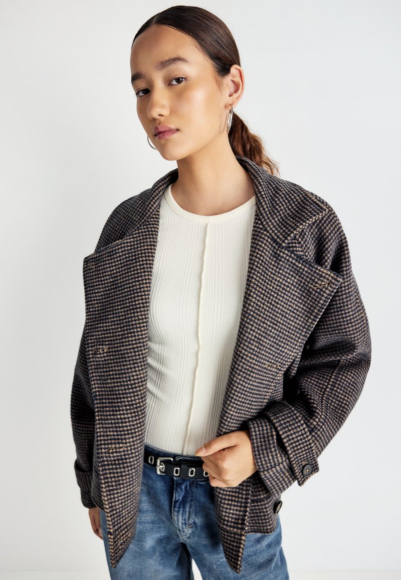 Ung kvinde med hestehale iført store hoops øreringe, en ternet oversized jakke, en hvid ribbet top og blå jeans med sort bælte.