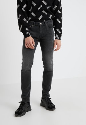 Man draagt slim-fit vervaagde zwarte jeans, zwart Versace Jeans Couture sweatshirt met witte logoprint en zwarte sneakers, staande tegen een witte achtergrond.