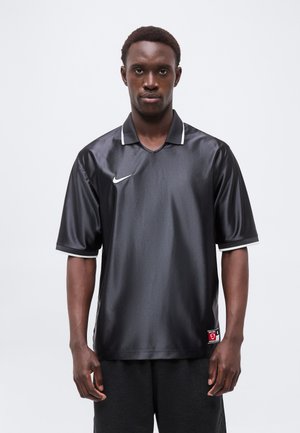 Junger Mann trägt ein glänzendes schwarzes Sporttrikot mit weißem Rand und weißem Nike-Logo, steht vor einem einfarbigen hellen Hintergrund.