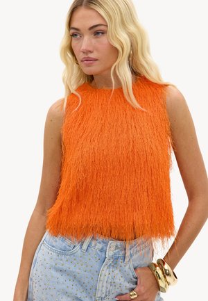 Femme blonde portant un haut sans manches texturé orange vif et un jean bleu clair avec des clous dorés, accessoirisée avec des bracelets épais en or et une bague.