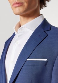 Blau strukturierte Anzugjacke mit Revers, weißem Taschentuch und einem weißen Button-Up-Hemd, das eine glatte Stoffoberfläche zeigt.