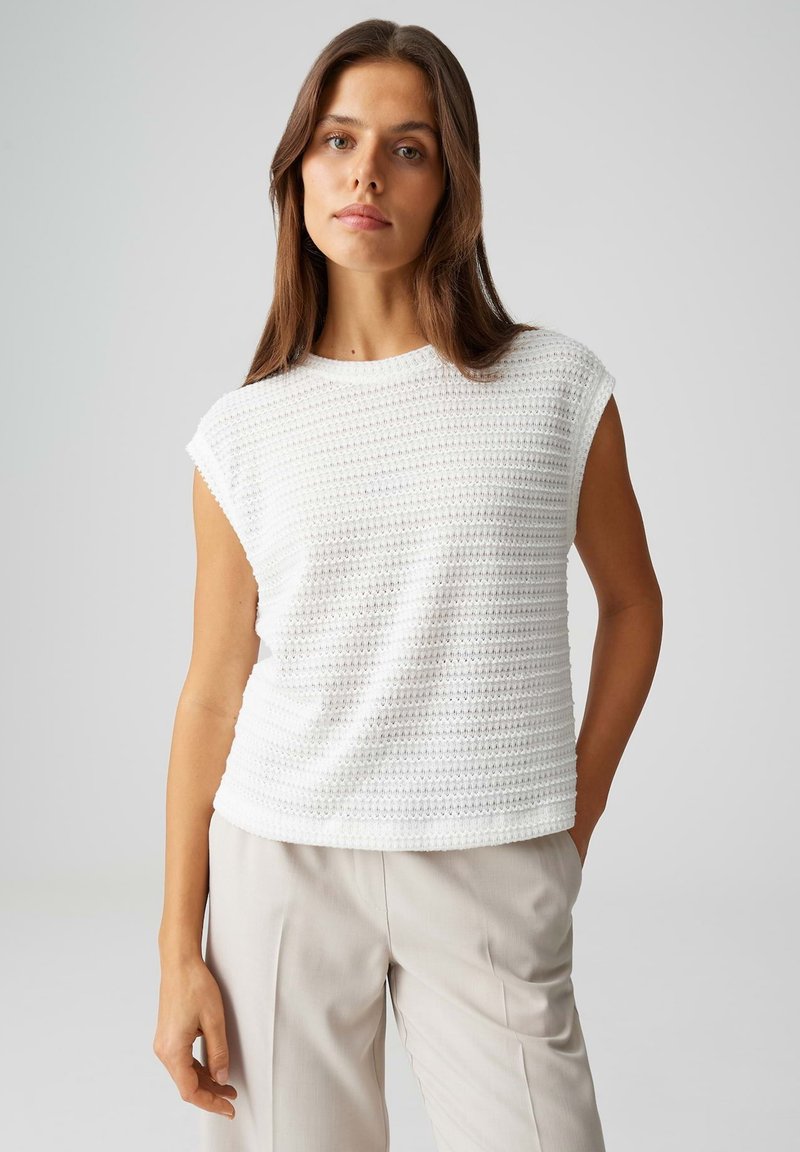 Donna con capelli castano lunghi che indossa un top bianco senza maniche con trama e pantaloni beige chiaro, in piedi davanti a uno sfondo grigio uniforme.