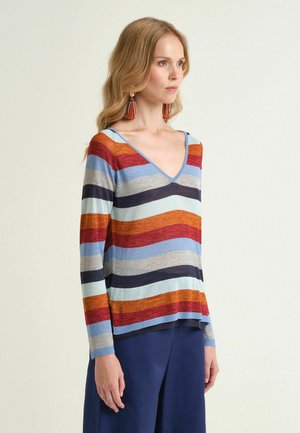 Donna che indossa un maglione a righe con scollo a V in blu, rosso, arancione, grigio e blu navy con maniche lunghe e pantaloni navy a gamba larga, rivolta verso destra.