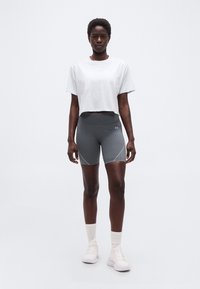 Γυναίκα όρθια, φοράει λευκό κοντό τοπ, γκρι ποδηλατικά σορτς ψηλής μέσης, λευκές κάλτσες και λευκά αθλητικά παπούτσια σε απλό φόντο.