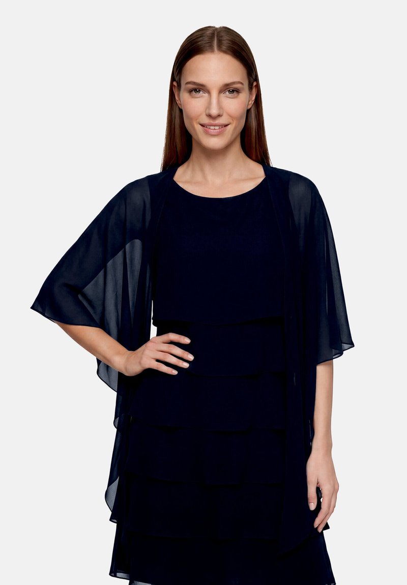 Frau mit glattem braunem Haar, die ein mehrlagiges dunkelblaues Kleid und einen transparenten dunkelblauen Schal trägt, steht mit einer Hand in der Hüfte und lächelt.