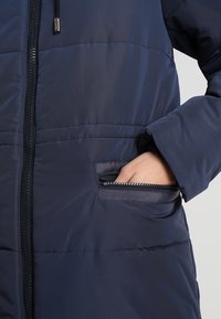 Chaqueta acolchada de color azul marino con una textura suave, que presenta un cierre frontal y un bolsillo con cremallera en ángulo en el lado izquierdo.
