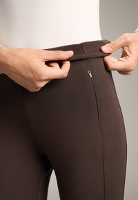 Bruine legging met een gladde textuur, uitgerust met een verborgen ritszak en een brede tailleband, die met de handen wordt aangepast.