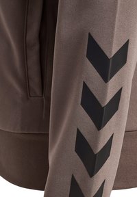 Brown jacket featuring a textured fabric and a ribbed hem, with black chevron patterns running down the left sleeve.