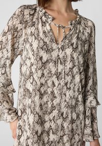 Bluzka z długim rękawem w wężowy wzór w odcieniach beżu i brązu, z dekoltem typu keyhole z wiązaniami oraz falbanianymi mankietami. Lekka, teksturowana tkanina.