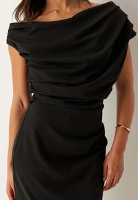 Zwarte jurk met gedrapeerde stof, asymmetrische halslijn en nauwsluitend silhouet. Soepele textuur met subtiele glans. Gouden armband als accent.