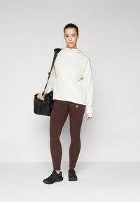 Sudadera con capucha de color crema claro, leggings marrones y zapatos negros, con una mochila deportiva negra y un pequeño logo en la parte superior. Textura suave y ajustada.