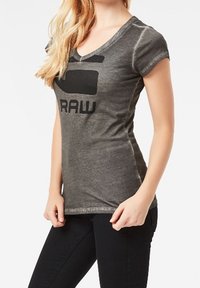 Camiseta gris ajustada de cuello en V, con mangas cortas, que presenta un gran logotipo negro "RAW" en el frente, hecha de una tela suave y texturizada.