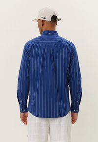 Camisa de manga larga azul con rayas verticales blancas, cuello abotonado y detalle de un plisado en la parte posterior. Presenta puños abotonados y una tela duradera.