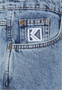 Šviesiai mėlynos džinsinės kelnės turi nugarinį kišenę su baltu išsiuvinėtu logotipu, vario praraitais ir matomu siuvimu. Šiek tiek nuvalyta tekstūra.