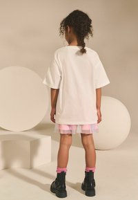Bílé oversized tričko položené na pastelově růžovou tylovou sukni, doplněné černými armádními botami a růžovými kotníkovými ponožkami. Pozadí obsahuje geometrické tvary.