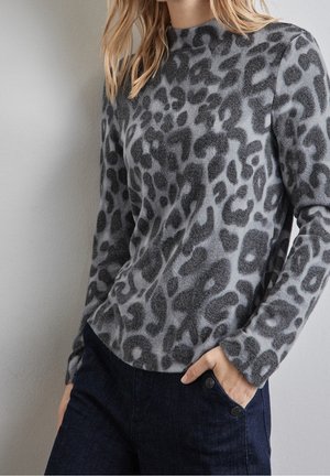 Frau trägt einen grau-schwarzen Leopardenmuster-Pullover mit langen Ärmeln und dunkelblaue Hose, eine Hand in der Tasche vor einem schlichten Wandhintergrund.