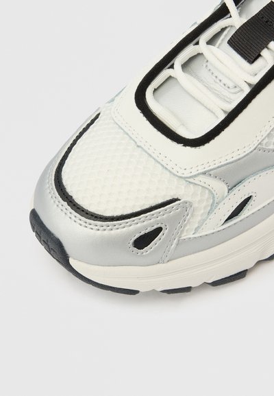 Λευκό αθλητικό παπούτσι με υλικά από δίχτυ και ασημένιο δέρμα. Διαθέτει μαύρες λεπτομέρειες, υφή στην επιφάνεια και παχιά, λαστιχένια σόλα με αυλακώσεις.