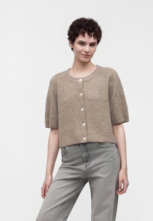 Jeune femme aux cheveux courts et bouclés portant un cardigan beige boutonné à manches courtes et un jean gris sur un fond uni.