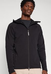 Crna zipped hoodie s pripijenim dizajnom, mekom teksturom i kontrastnim šavovima. Sadrži prednji zip i bočne džepove.