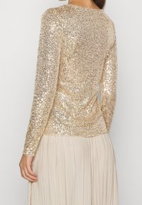 Langarmtop in gouden pailletten met een gestructureerde afwerking. Kenmerken omvatten een ronde halslijn en een aansluitende pasvorm, gecombineerd met een geplooide beige rok.