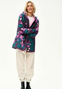 Kobieta uśmiechająca się, ze skrzyżowanymi ramionami, ubrana w fioletową kurtkę z dużymi turkusowymi i białymi kropkami, białe spodnie oraz czarne botki za kostkę.