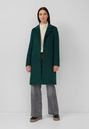 Manteau long en mélange de laine vert avec des revers crantés, trois boutons et poches latérales, associé à un pull crème et un jean gris évasé.