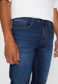 Dark blue denim jeans with a slim fit, featuring a classic five-pocket design and contrasting stitching. The front pocket shows a red accent.