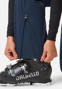 Hände ziehen den Reißverschluss einer dunkelblauen Hose über einem schwarzen Dalbello-Skieboot mit Metallriegelschnallen vor einem weißen Hintergrund hoch.