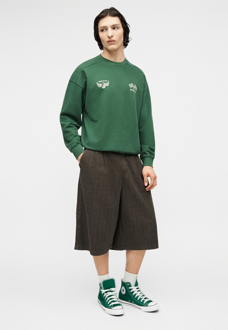 Jeune personne portant un sweat-shirt vert avec des logos blancs, un short ample brun foncé jusqu'aux genoux, des chaussettes blanches et des baskets montantes vertes, immobile.