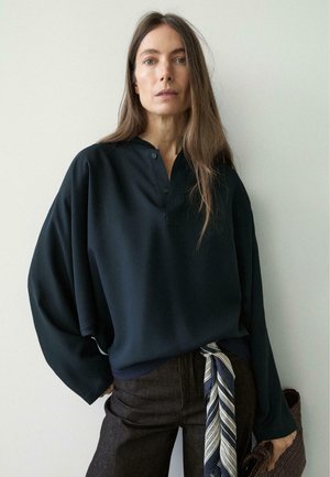 Femme aux longs cheveux bruns portant une chemise noire ample et un pantalon foncé avec une ceinture écharpe rayée, tenant un sac tissé sur un fond uni.