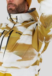 Gul og hvid mønstret jakke med lynlåslukning foran, justerbar hætte og sorte hardware-detaljer. Har et glat, tekstureret stof.