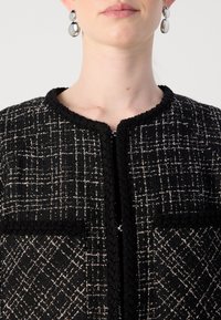 Femme portant une veste texturée à carreaux noirs et blancs avec un col rond et des boucles d'oreilles pendantes en argent.