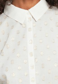 Biała bluzka z kołnierzykiem na guziki, ozdobiona jasnym złotym wzorem w kropki oraz teksturowanym materiałem.