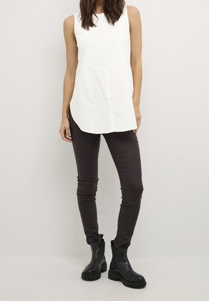 Nainen, joka käyttää hihattoman valkoisen napillisen tunikapaidan, tummat kapeat housut ja mustat massiiviset nilkkurit, seisoo yksinkertaisen vaalean taustan edessä.