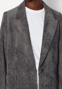 Blazer grigio in corduroy con texture a righe verticali, rever a punta, chiusura con un solo bottone e due tasche frontali, indossato su una camicia bianca.