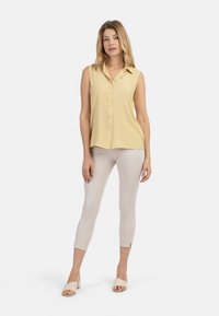 Lichtgele mouwloze blouse met een kraag en knopen aan de voorkant, gecombineerd met lichtbeige aangesloten pantalons en zachte nude blokhakken sandalen.