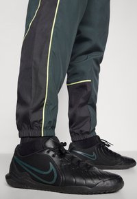 Czarne sportowe buty Nike z teksturowaną powierzchnią i zielonymi akcentami, zestawione z ciemnymi spodniami sportowymi z kontrastowymi czarno-żółtymi paskami.