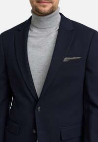 Marineblauwe blazer van een gestructureerde stof, met twee knopen en zijzakken, gedragen over een lichtgrijze turtleneck sweater.