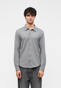 Πουκάμισο με μακριά μανίκια και κουμπιά σε ανοιχτό γκρι χρώμα, κατασκευασμένο από μαλακό βαμβακερό ύφασμα. Διαθέτει κλασικό γιακά και καμπυλωτό τελείωμα. Φορεμένο με σκούρο τζιν.