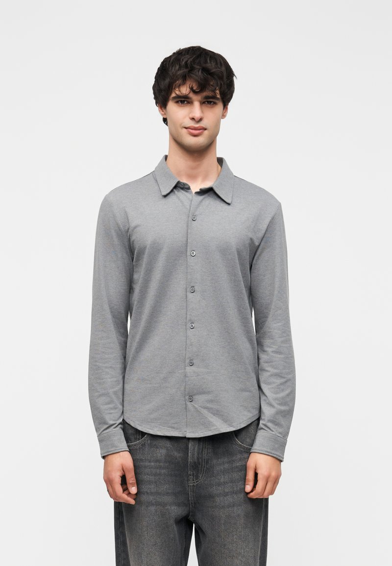 Πουκάμισο με μακριά μανίκια και κουμπιά σε ανοιχτό γκρι χρώμα, κατασκευασμένο από μαλακό βαμβακερό ύφασμα. Διαθέτει κλασικό γιακά και καμπυλωτό τελείωμα. Φορεμένο με σκούρο τζιν.