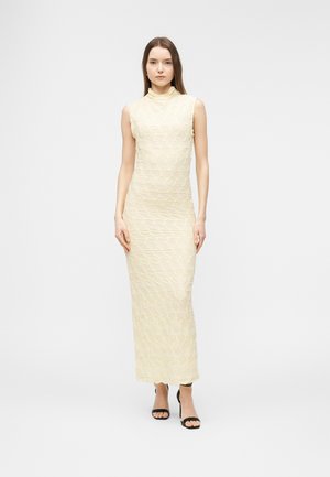 Kobieta stojąca w bezrękawowej kremowej sukience o fakturze, sięgającej kostek, z wysokim kołnierzem i czarnych, odkrytych na palcach szpilkach, na jednolitym tle.