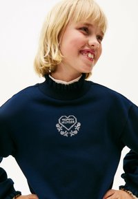 Námornícky sveter s volánovým golierom, s bielym vyšitým srdcom s nápisom "MOMMY HILFIGER" a kvetinovými akcentmi. Mäkká textília.