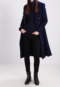 Marineblauer Wollmantel mit Doppelreiher-Design und silbernen Knöpfen. Kombiniert mit einem marineblauen Grobstrickpullover, schwarzen Skinny-Hosen und Ankle Boots.