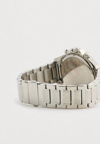 Ezüst színű fémóra, láncos karkötővel és csattal, a hátsó tokot és állító gombokat mutatja, sima világos háttér előtt.